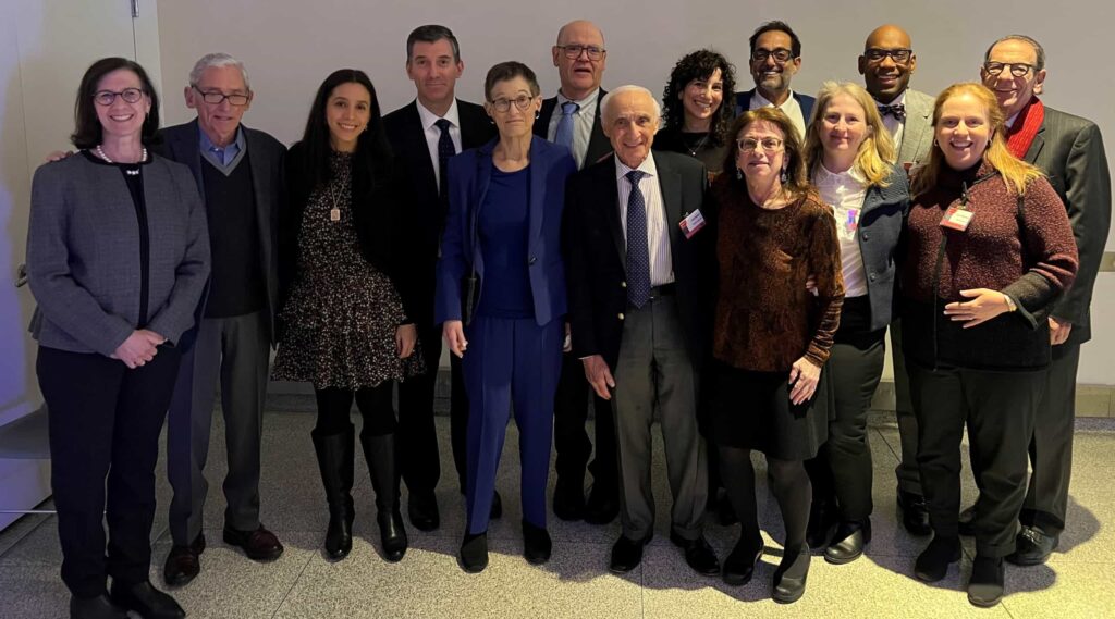 A diverse group of thirteen individuals, dressed in business or semi-formal attire, stand together indoors at Temple Emanu-El in New York City, smiling warmly at the camera. The group includes both men and women of various ages, gathered against a softly lit background.
