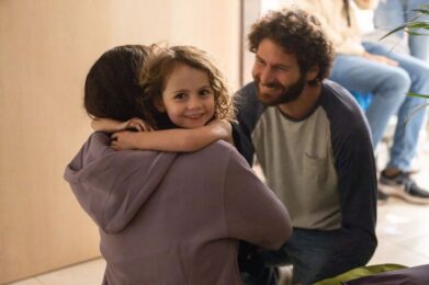 A joyful young girl with curly hair embraces an adult wearing a purple hoodie, as another adult with a beard smiles warmly nearby. The group is gathered indoors at Temple Emanu-El, surrounded by fellow community members.