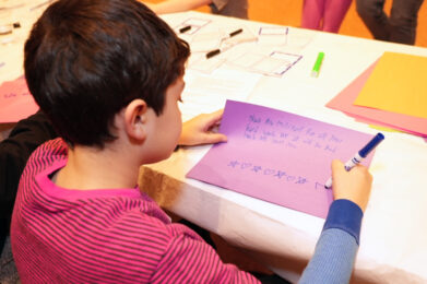 A young child participates in a creative activity at a table filled with colorful craft supplies and papers, joined by others in a warm, communal setting.