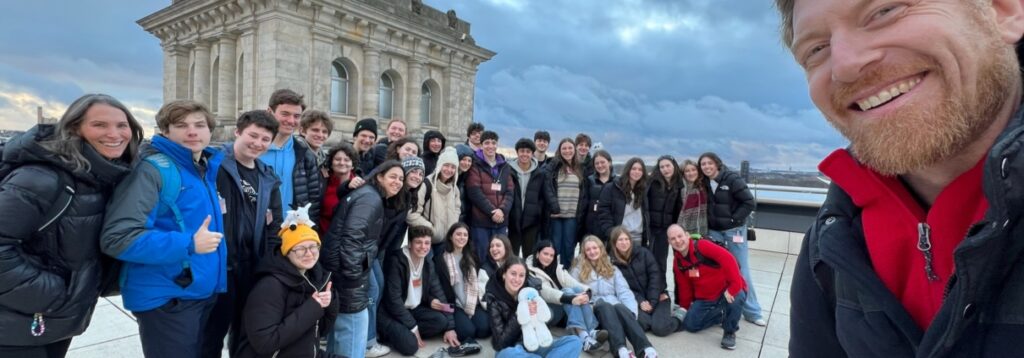 A joyful group of Temple Emanu-El teens and adult chaperones gather for a rooftop photo, with a beautiful historic stone building and cloudy sky in the background. One participant takes the selfie from the front.