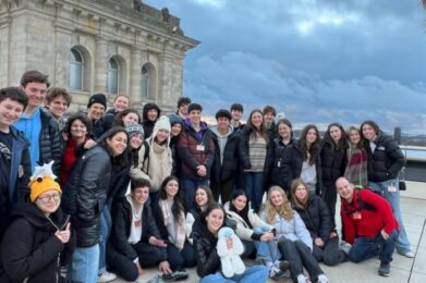 A joyful group of Temple Emanu-El teens and adult chaperones gather for a rooftop photo, with a beautiful historic stone building and cloudy sky in the background. One participant takes the selfie from the front.