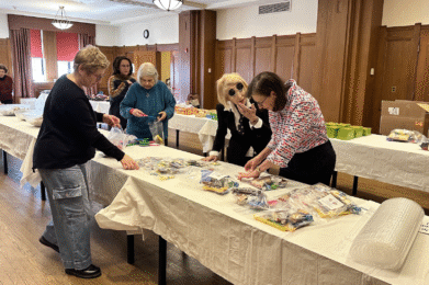 A group of women from Temple Emanu-El gather around tables in a bright, welcoming room with wooden paneling and large windows, assembling care packages to support those in need.