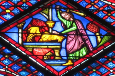 A stained glass window illustrates the biblical story of Abraham and Isaac, showing Abraham, crowned and holding a raised arm, standing over Isaac on an altar, framed by vibrant red, blue, and yellow geometric patterns.