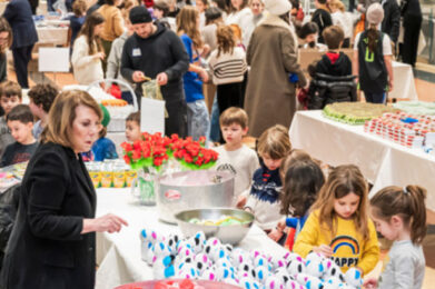 A large group of children and adults gather indoors around tables covered with colorful craft supplies and snacks, participating in a lively community event at Temple Emanu-El.