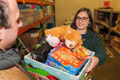 A smiling volunteer from Temple Emanu-El hands a box filled with toys, such as a teddy bear and board games, to another person in a storage area lined with shelves of donated items.