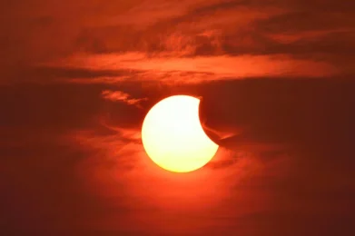 A partially eclipsed sun shines with warm orange and yellow hues through a cloud-filled, softly red-tinted sky, as the moon gently covers part of the sun’s upper right edge.