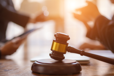 A wooden gavel rests on a desk in the foreground, while people in formal attire engage in conversation over documents in the background, evoking themes of justice and ethical discussion within a communal setting.