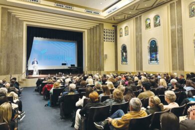 A large audience gathers in a beautifully decorated sanctuary with stained glass windows, watching a presenter speak onstage near a grand piano and a large screen displaying the “YESOD” logo at Temple Emanu-El.