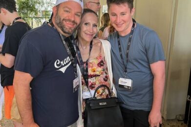 Three members of the Temple Emanu-El community stand together, smiling for a photo during a synagogue event. All are wearing event lanyards; one holds a shopping bag, another carries a black purse. Other participants can be seen in the background.
