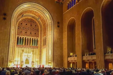A large congregation gathered inside Temple Emanu-El's majestic Fifth Avenue Sanctuary with soaring arched ceilings, intricate decorative details, and tall stained glass windows illuminated by warm lighting.