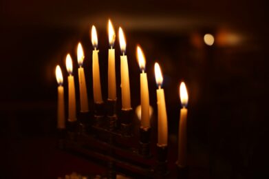 A fully lit menorah with nine candles shining brightly against a dark background, representing the celebration of Hanukkah at Temple Emanu-El.