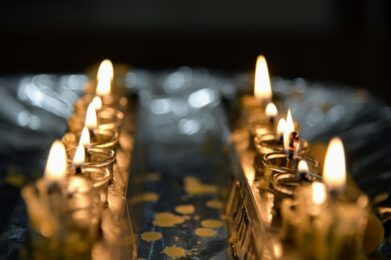 Two rows of small Hanukkah oil lamps with bright flames are arranged on a reflective surface, their warm glow symbolizing the miracle of Hanukkah. The background is softly blurred to highlight the festive lights.