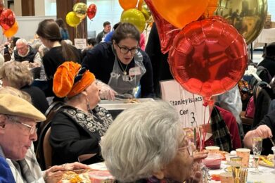 A joyful group of seniors share a meal together at a beautifully set table adorned with festive balloons at Temple Emanu-El's annual Thanksgiving Dinner, which is hosted by the temple's Tikkun Olam Committee for people from senior centers, shelters for people facing housing insecurity, and shelters protecting women and childreni from domestic violence.