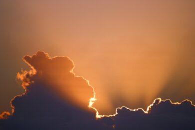The sun sets behind gentle clouds, casting a warm glow of orange and gold across the evening sky above Temple Emanu-El, evoking a sense of tranquility and reflection.