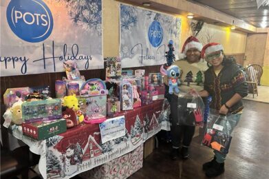 Two people in festive hats smile beside a display of toys and gifts donated by Temple Emanu-El members on a decorated table, with banners reading Happy Holidays in the background.