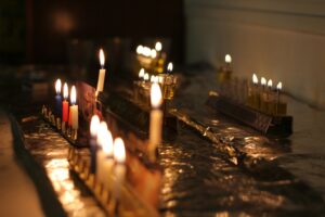 A close-up image of several hanukkiyot (Hanukkah menorahs) each glowing with colorful candles and oil flames, arranged together on a foil-covered table. The warm candlelight creates a joyful and inviting Hanukkah celebration scene.