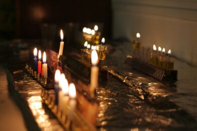 A close-up image of several hanukkiyot (Hanukkah menorahs) each glowing with colorful candles and oil flames, arranged together on a foil-covered table. The warm candlelight creates a joyful and inviting Hanukkah celebration scene.
