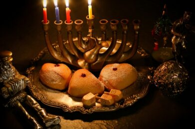 A Hanukkah menorah (hanukkiyah) with six candles aglow stands behind a silver tray displaying three sufganiyot (jelly-filled donuts), along with dreidels and decorative holiday figurines, all beautifully arranged on a dark tabletop.