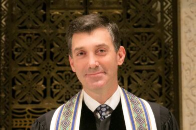 A clergy member wearing formal robes and a decorative tallit stands in front of an ornate, gold-patterned background, smiling warmly at the camera.