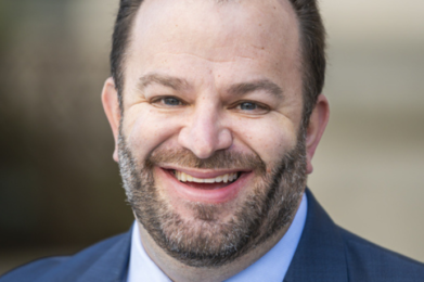 A smiling man with short brown hair and a beard, dressed in a blue suit, light blue shirt, and blue tie, stands outside against a softly blurred background.