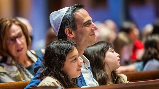 A person wearing a white kippah sits with two children in a crowded room at Temple Emanu-El, all facing forward and appearing engaged in a communal gathering or service.