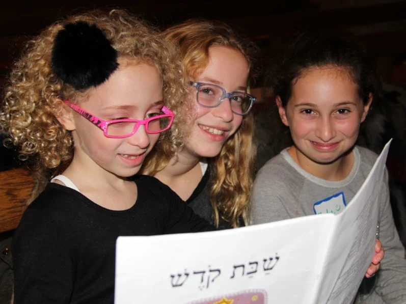 Three smiling girls sit together at Temple Emanu-El. One, with curly blonde hair and pink glasses, holds a paper featuring Hebrew text as the other two lean in to look, all sharing a joyful moment of learning.