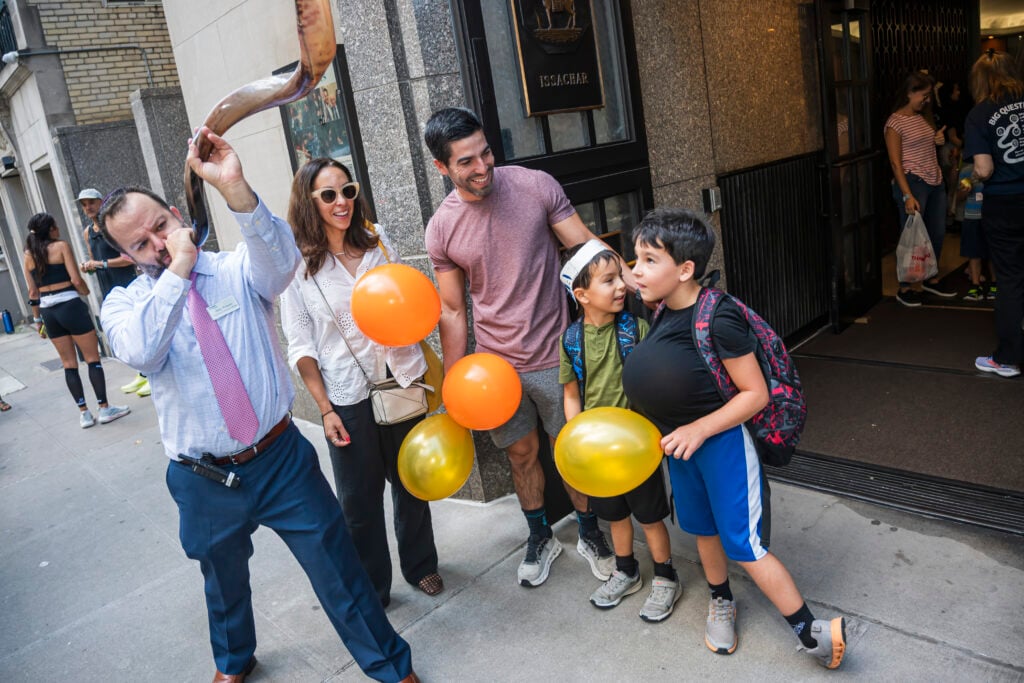 A man sounds the shofar on a New York City sidewalk as families from Temple Emanu-El, including children with balloons, look on.