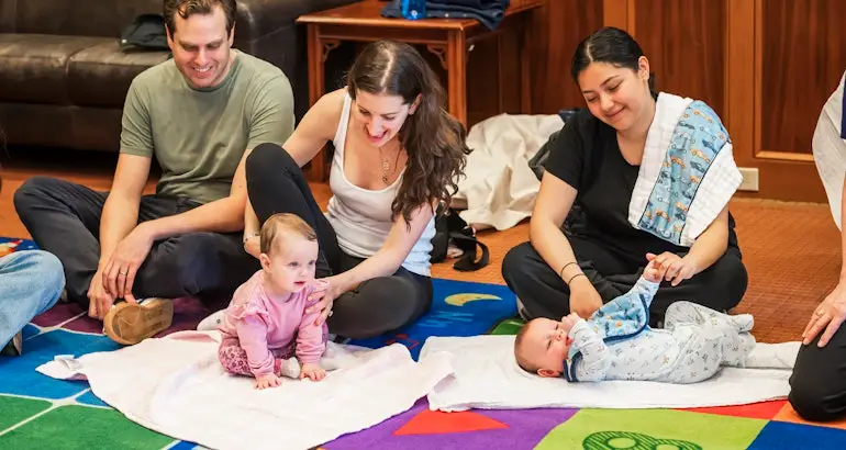 Three adults gather on a colorful mat with two babies; one baby crawls while the other rests on their back, sharing joyful moments.