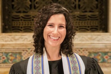 A woman with curly brown hair, wearing a decorative tallit over formal attire, smiles while standing indoors in front of an ornate synagogue backdrop.
