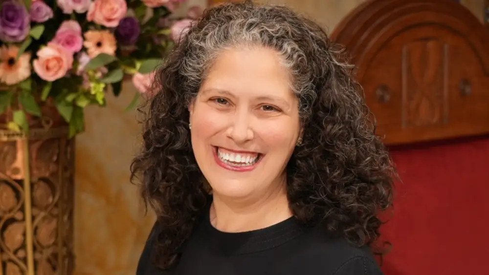 Smiling woman with curly hair seated near floral arrangements and a wooden chair with a red cushion at Temple Emanu-El.