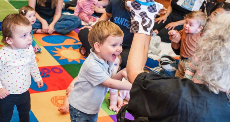 A group of young children gathers on a colorful play mat in a bright room at Temple Emanu-El, joyfully watching an adult lead them in an engaging puppet activity. The children look curious and delighted as they participate together.
