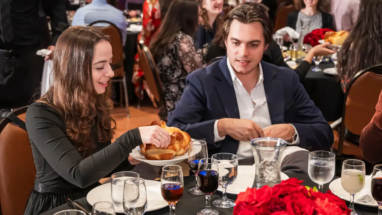 A young woman and man sit at a round table set for a formal meal at Temple Emanu-El, with the woman passing challah. The table is arranged with glasses, plates, and a centerpiece featuring red flowers. Other congregants are dining and conversing in the background.