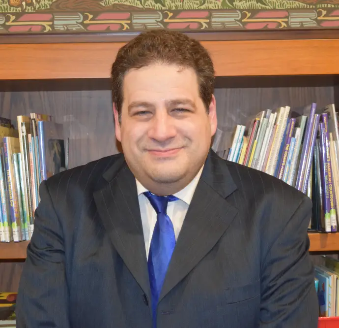 A man in a suit and blue tie smiles warmly in front of a bookshelf, standing in a welcoming space at Temple Emanu-El.