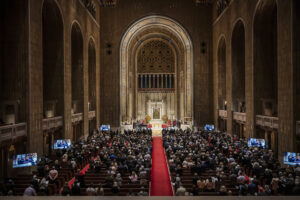A large audience sits in pews facing the ornate bimah of a grand, high-ceilinged sanctuary with arched architecture and a red carpet down the center aisle.