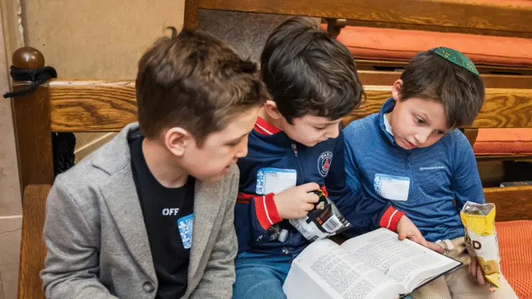 Three children sit together on a wooden bench at Temple Emanu-El, reading and sharing an open book. They look engaged and are dressed casually with name tags, with snacks nearby.