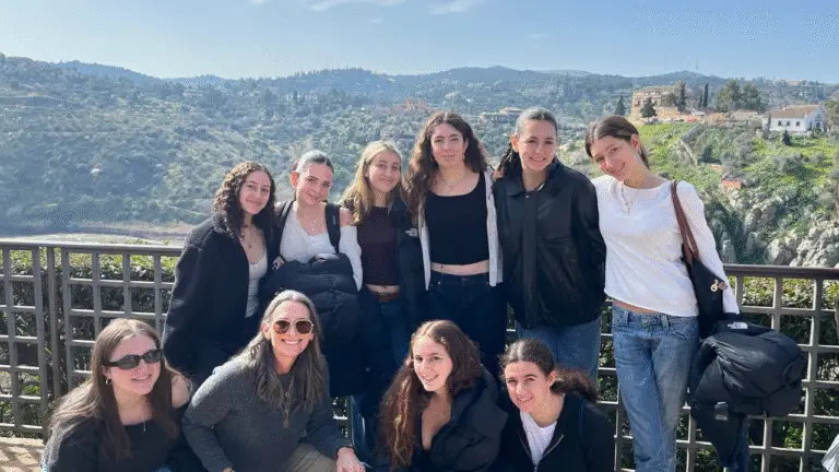 A group of ten young women, members of the Temple Emanu-El community, gather and smile together outdoors in front of a scenic landscape with green hills, trees, and blue sky. They are dressed casually for the occasion.