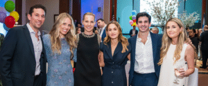 Six adults, members of the congregation, gather and smile at a Temple Emanu-El social event. They are dressed in semi-formal attire, with festive decorations and other attendees visible in the background. One woman holds a glass during the celebration.