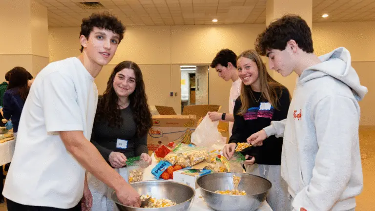 Five teenagers gather around a table at Temple Emanu-El, smiling as they prepare snack bags for a community service project. They work together, scooping food from large bowls, with boxes and bags of snacks visible in the background.