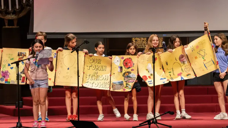 A group of children stand on the bimah holding a long, illustrated banner that reads Our Torah, Jewish Stories. One child speaks into a microphone while others proudly display the colorful artwork.