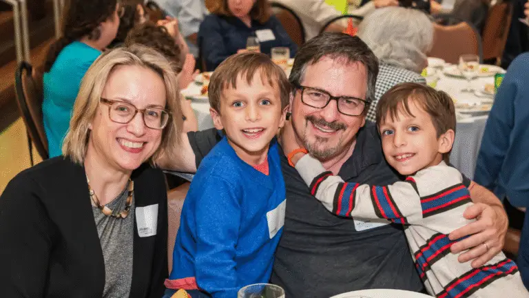 A smiling family of four gathers together at a shared table during a Temple Emanu-El community event. The parents and their two young sons sit closely, expressing warmth and joy as they celebrate together.
