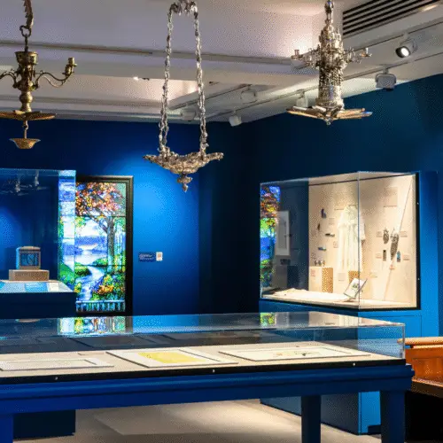 A museum exhibit with blue walls showcases stunning stained glass art, elegant silver chandeliers, and display cases containing Jewish artifacts and historical documents. A wall-mounted text panel offers context for visitors. Spotlights highlight the vibrant colors of the glasswork and items on display, creating an inviting atmosphere for learning about Jewish heritage.