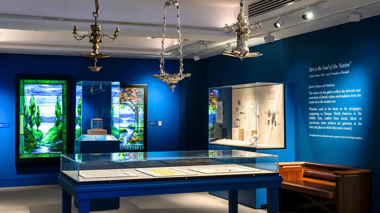A museum exhibit with blue walls showcases stunning stained glass art, elegant silver chandeliers, and display cases containing Jewish artifacts and historical documents. A wall-mounted text panel offers context for visitors. Spotlights highlight the vibrant colors of the glasswork and items on display, creating an inviting atmosphere for learning about Jewish heritage.