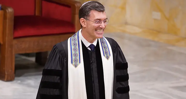 Rabbi Davidson wearing a tallit stands indoors near a large wooden chair, in the sanctuary of Temple Emanu-El.