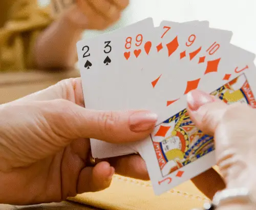 A person participates in a friendly card game at a table with others, holding a fan of playing cards.