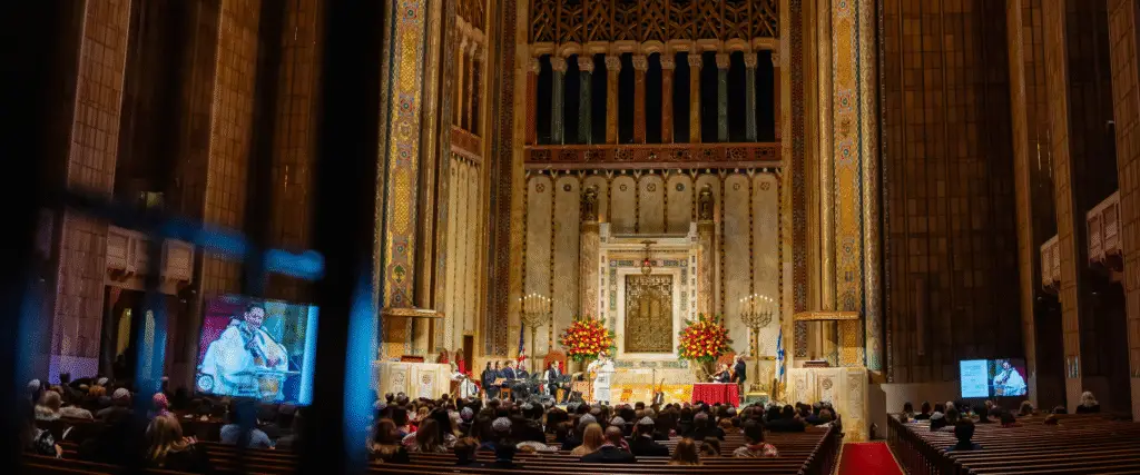 A large audience gathers inside Temple Emanu-El’s magnificent sanctuary, attentively listening to a panel discussion framed by tall columns and elegant floral arrangements.