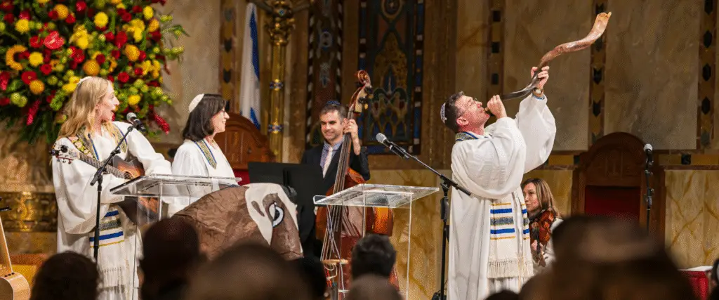 Five individuals in white robes participate in a musical performance inside Temple Emanu-El; one person blows a shofar while others play string instruments, contributing to the spiritual atmosphere of the synagogue.
