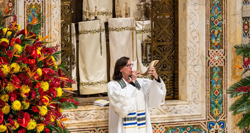 Individual in a white robe sounds the shofar before Torah scrolls and flowers at Temple Emanu-El in New York City.