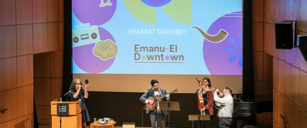 Four musicians perform on stage with Shabbat Shalom! Emanu-El Downtown projected behind them during a musical service at Temple Emanu-El.