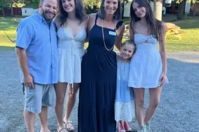 A smiling multigenerational family gathers outdoors, dressed in casual summer attire, with trees and picnic tables behind them, enjoying a warm day together.