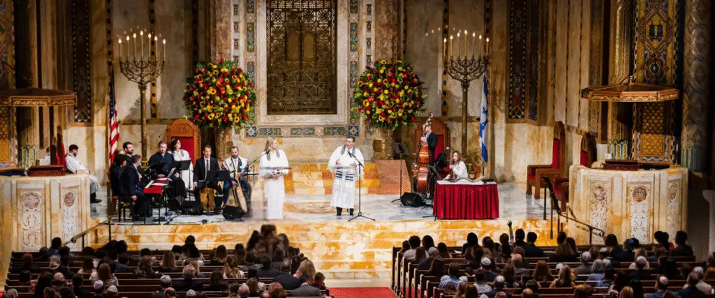 A vibrant community gathering at Temple Emanu-El, featuring musicians and speakers leading a service on the bimah in the synagogue’s grand sanctuary, with congregants seated in attendance.